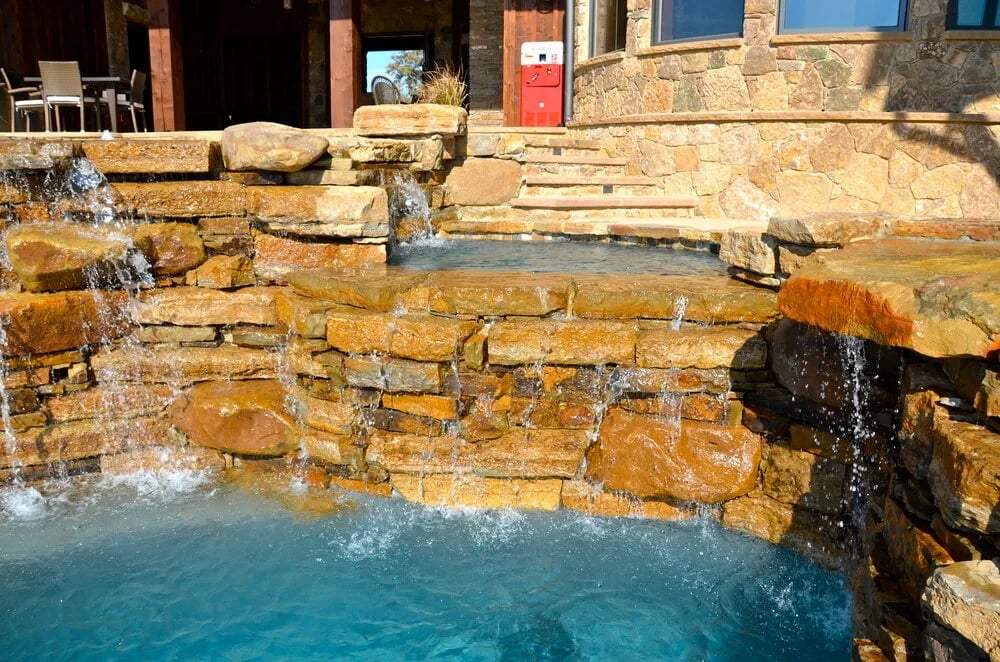 Layered stone waterfalls spilling into a sunlit freeform pool below a raised stone deck