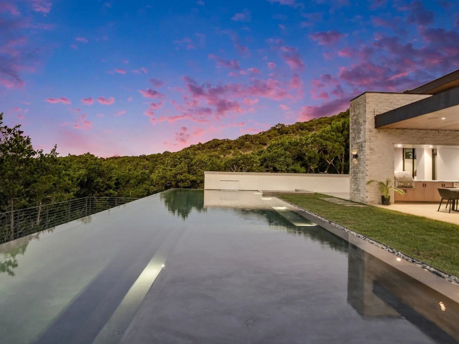 Modern stone-and-glass home glowing at dusk above an infinity pool and stone deck under a pink sky