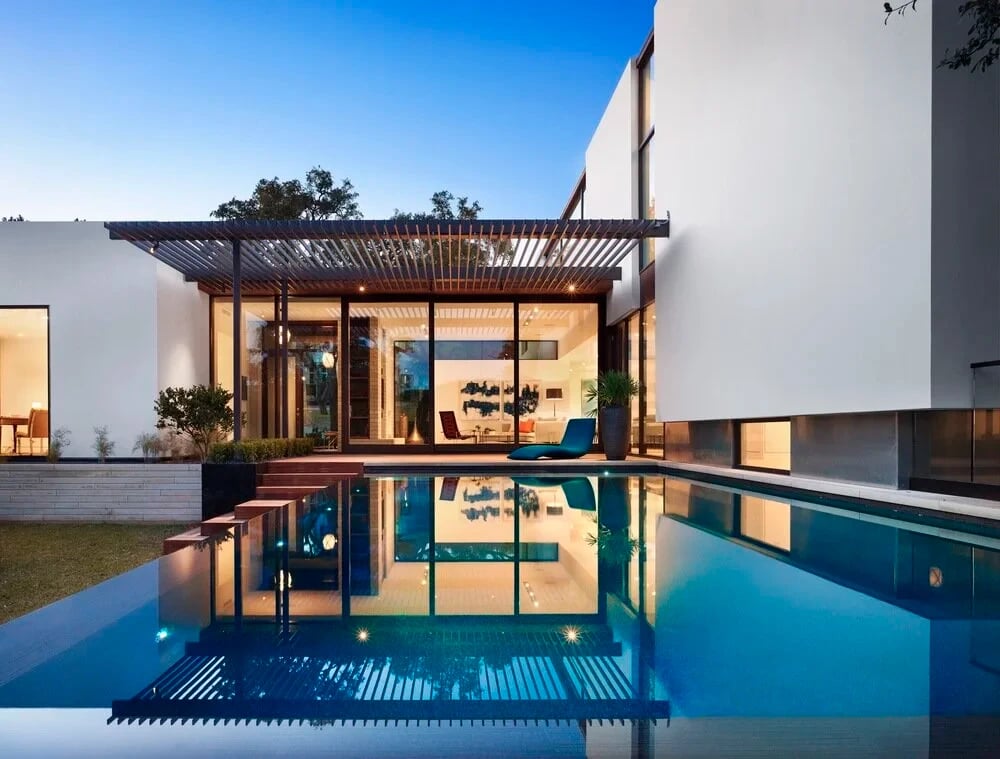 Infinity pool reflecting a lit white modern home and pergola at twilight, wood steps to the water