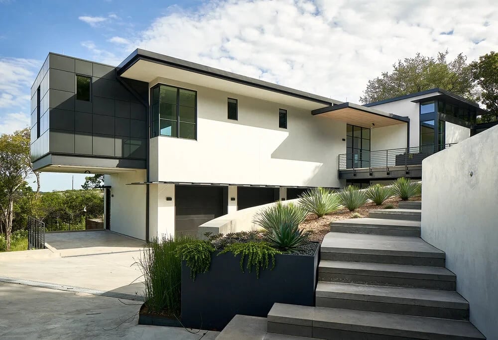 Two-story modern home with dark panels and white stucco above a carport, stairs and agave plantings