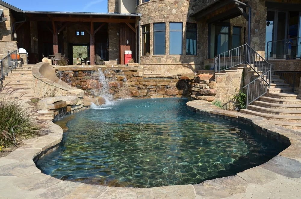 Freeform pool with a pebble interior and stone waterfall in a courtyard beside a stone home and steps
