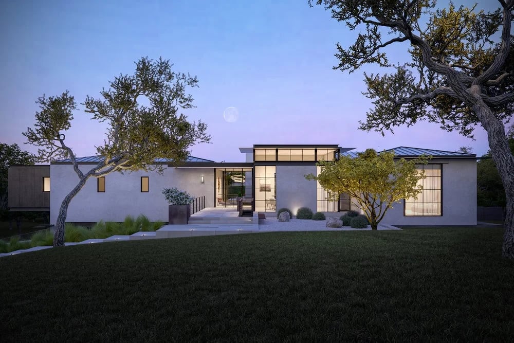 Low modern home with glass walls and a flat roof on a wide lawn at dusk, framed by oaks