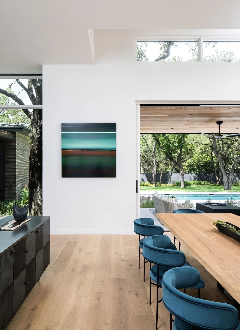 Dining nook with blue chairs and seascape art, glass walls opening to the backyard pool and trees