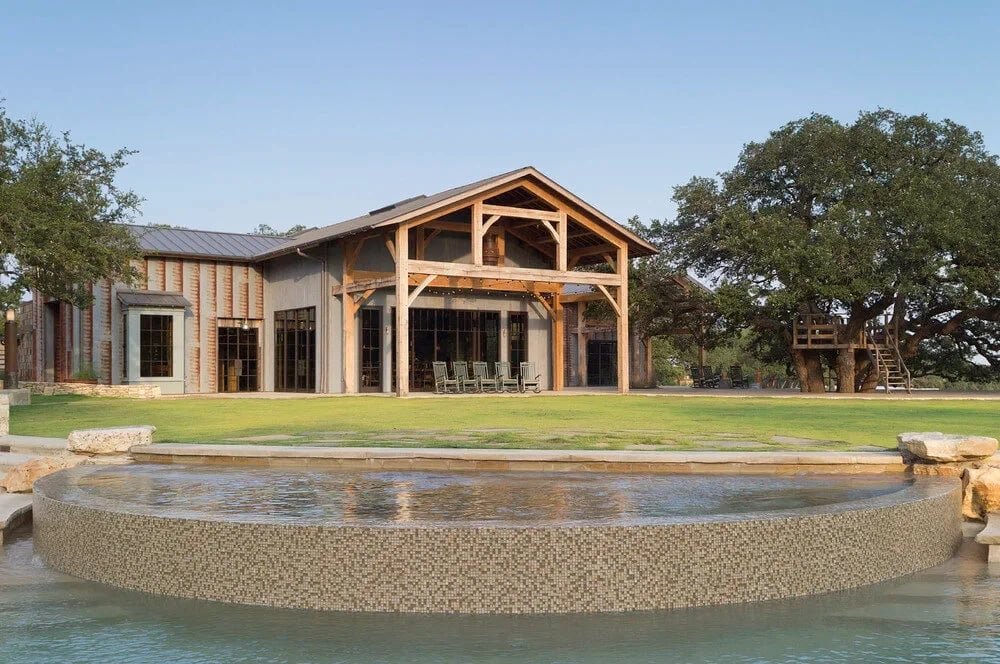 Timber-framed lodge with covered porch behind a still pebble-edged pool, framed by oaks